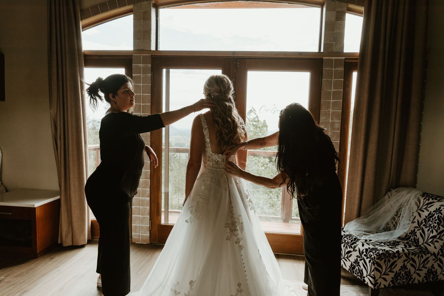 Two bridesmaids in black help a bride in a white lace gown prepare near a large window with mountain views.