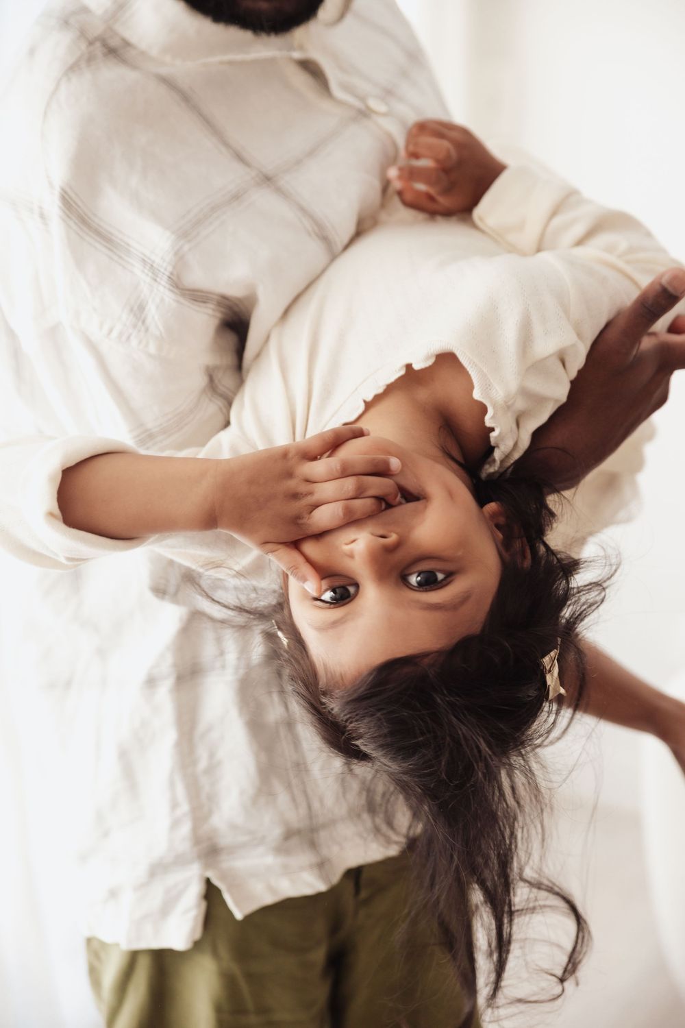 A child being held upside down by a parent, laughing and looking at the camera in a bright white room.