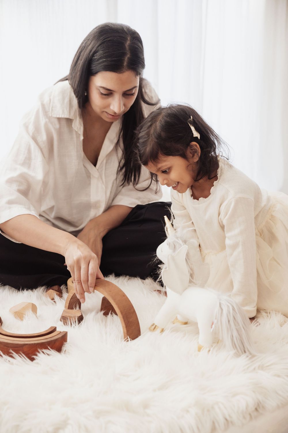 A mother and young daughter sit on a fluffy white rug playing with wooden toys and a white stuffed animal together.