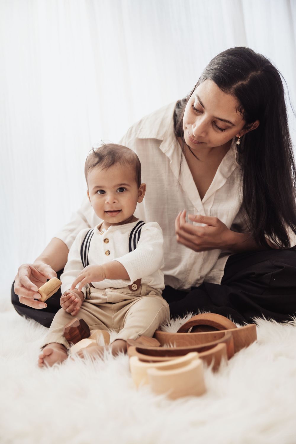 A mother and baby boy sit on a white fur rug playing with wooden toys together.
