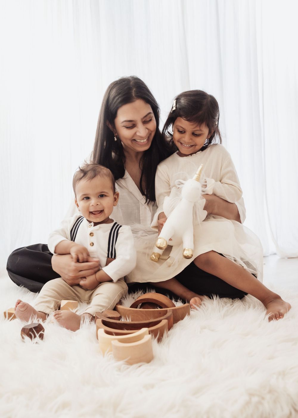A smiling mother sits on a fluffy white rug with her two young children, holding toys in a bright, airy room.