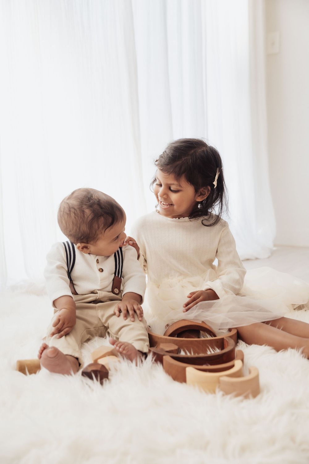 Two young children sit on a fluffy white rug playing with wooden toys near a bright window.