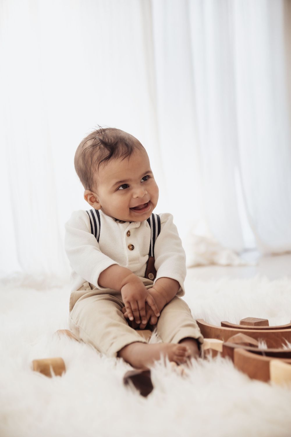 Adorable smiling baby boy in white shirt with suspenders sitting on fluffy rug with wooden toys.