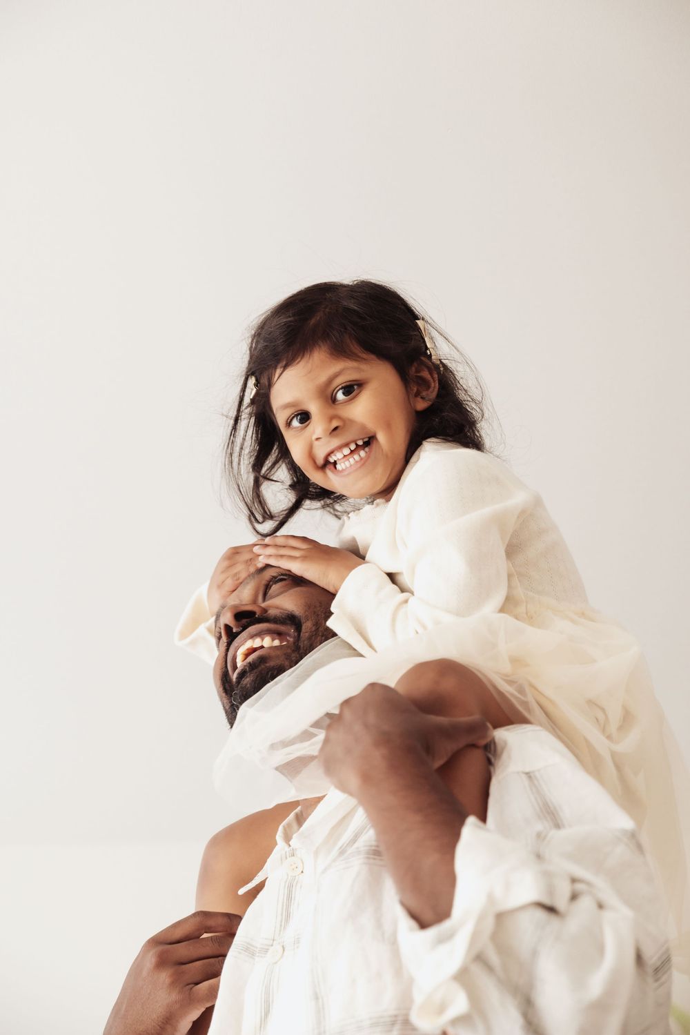 A joyful young girl in white dress rides on her smiling father's shoulders against a light background.