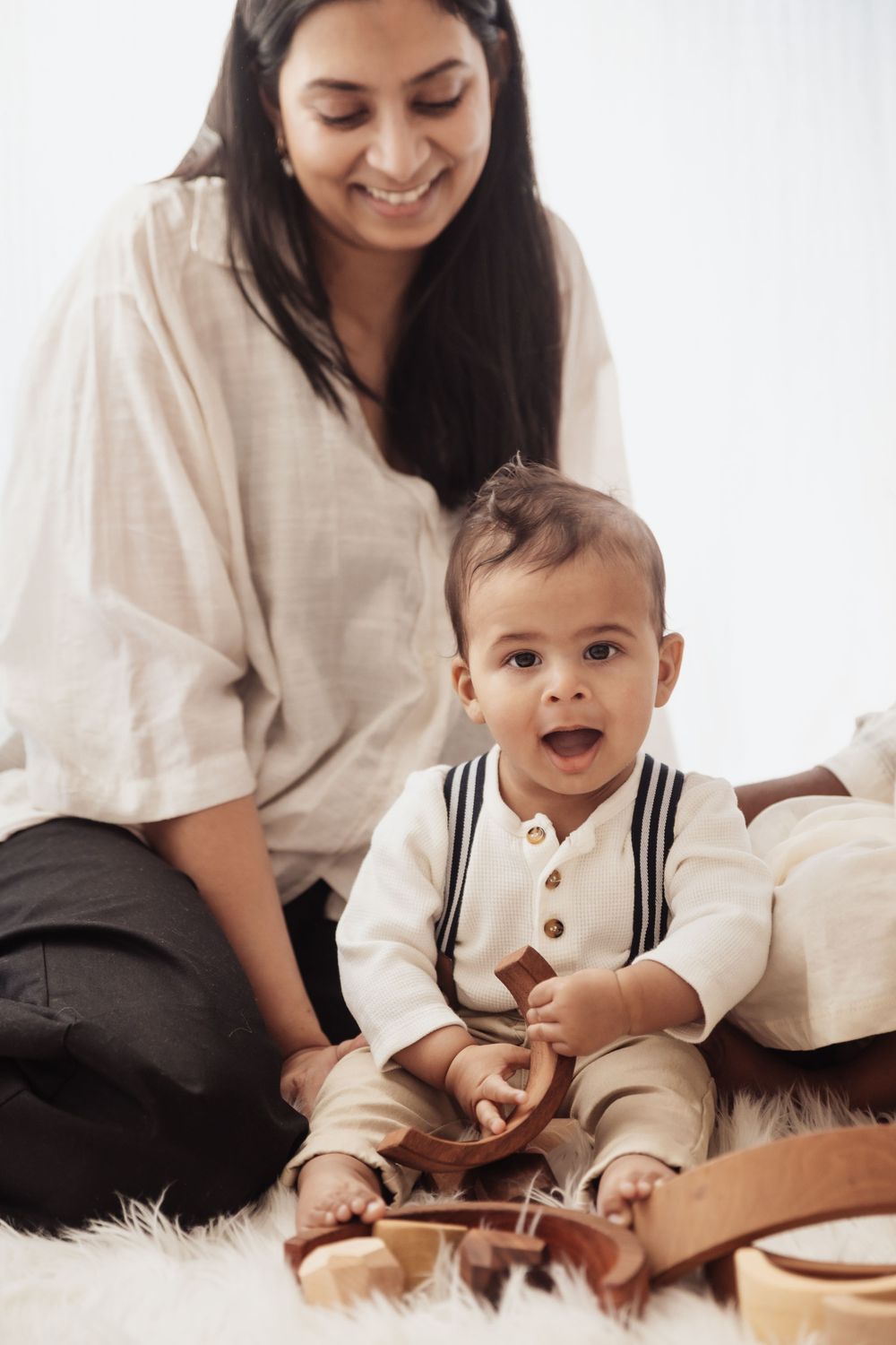 A smiling mother watches her happy baby boy play with wooden toys on a white fur rug.