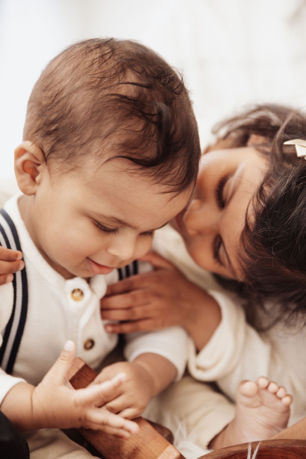A young girl smiles warmly while cuddling a baby in white clothing in a bright, cozy setting.