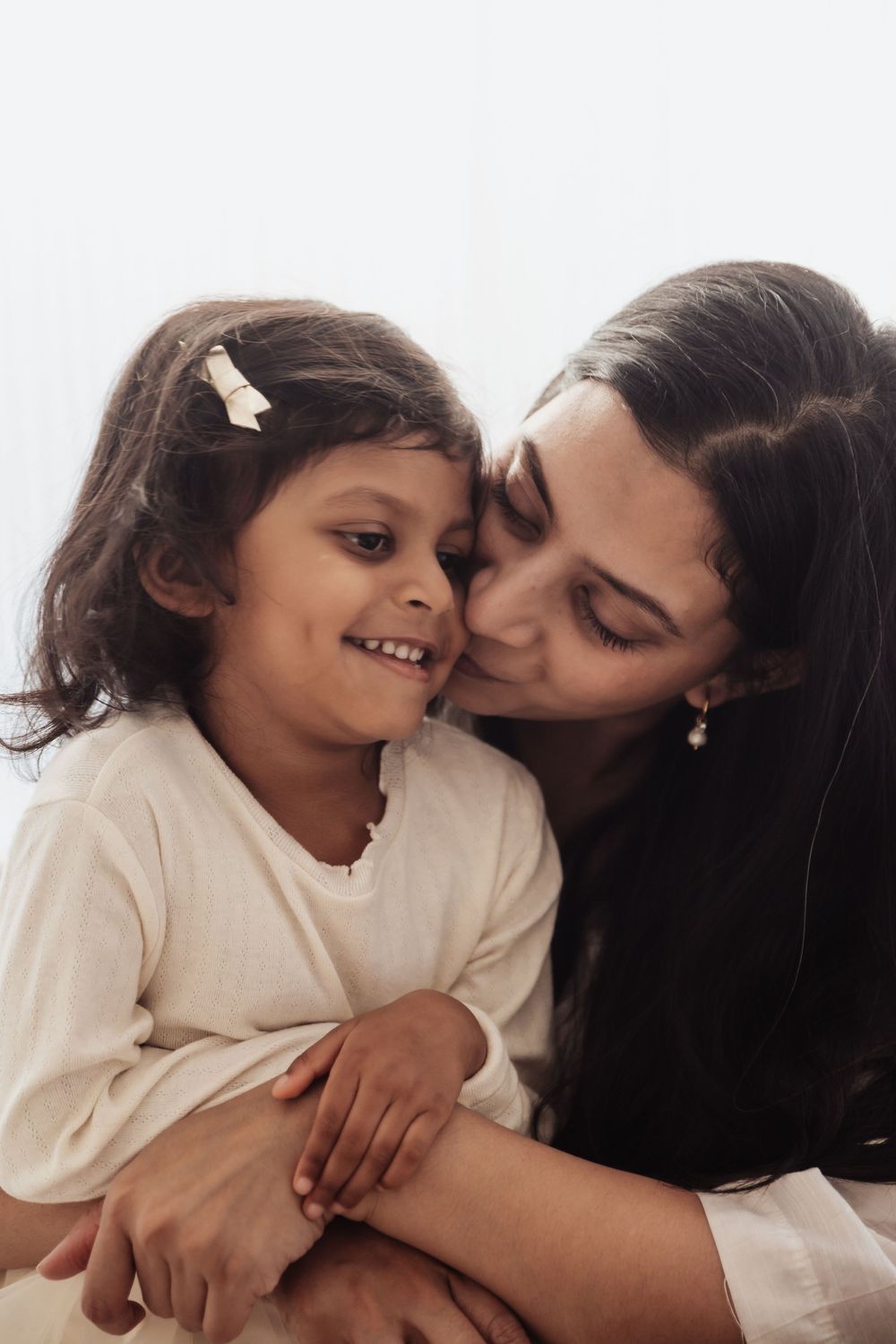 A woman tenderly kisses a smiling young girl's cheek while embracing her in a warm, heartfelt moment.