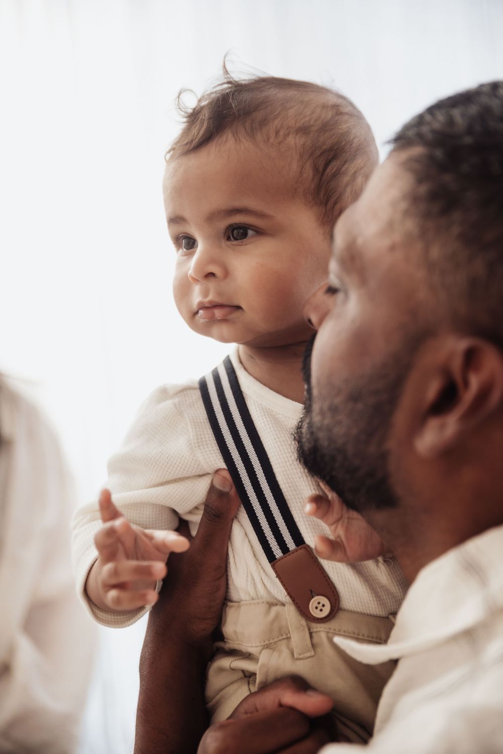 A bearded father lovingly holds his baby dressed in suspenders near a bright window.
