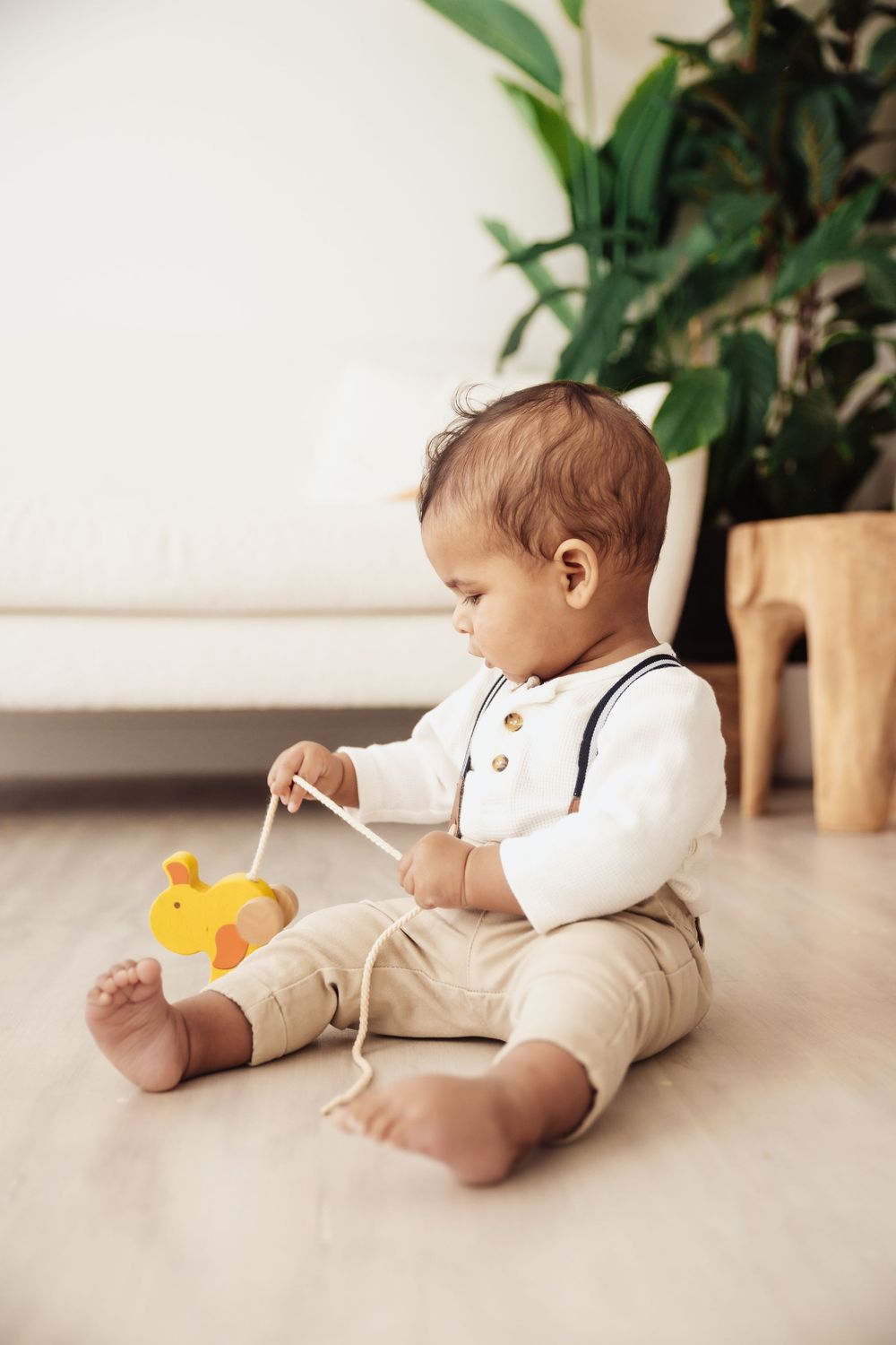 A cute baby in suspenders sits on the floor playing with a yellow wooden duck pull toy.
