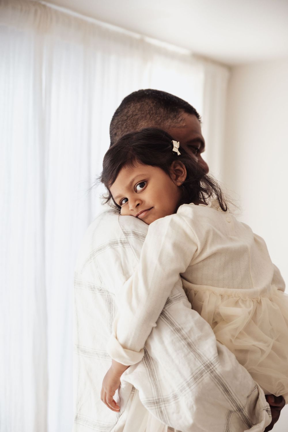 A young girl rests her head on a man's shoulder as he holds her in a warm embrace near a bright window.
