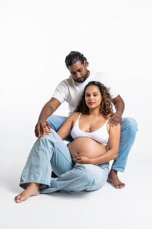 Pregnant woman in white bra and jeans sits with partner behind her, both on white background.