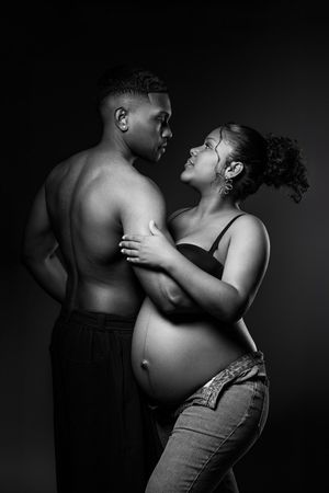 Black and white maternity photo of a pregnant couple embracing intimately against a dark studio background.