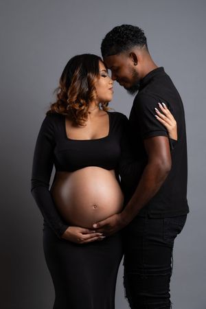 A pregnant Black couple in matching black outfits share a tender moment, foreheads touching, cradling her baby bump.