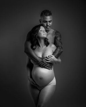 Black and white maternity photo of pregnant woman embraced by tattooed partner in intimate studio portrait.