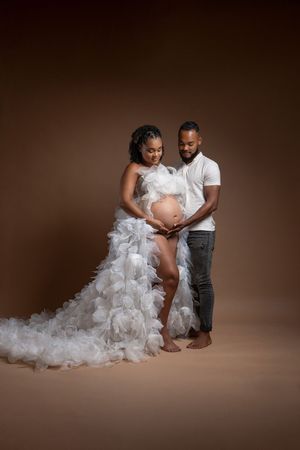 Couple in maternity photoshoot, pregnant woman in white ruffled gown, partner embracing her belly on brown background.