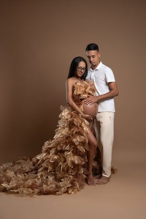 Pregnant woman in gold ruffled gown with partner in white outfit posing for maternity photo on brown background.