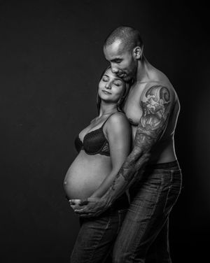 Black and white maternity photo of a tattooed shirtless man embracing a pregnant woman in a studio setting.