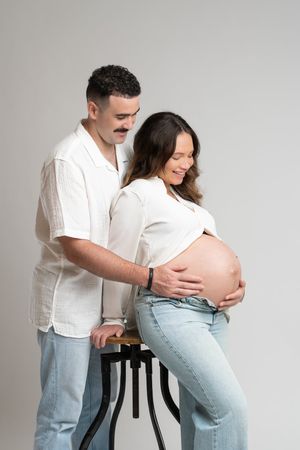Couple in white shirts and jeans posing in studio, pregnant woman cradling baby bump while partner stands behind her.