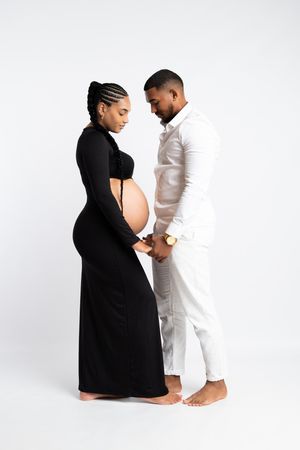 A pregnant couple in a studio, woman in black dress, man in white outfit, holding hands and looking at her baby bump.