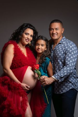 Pregnant woman in red tulle robe poses with her daughter holding a rose and husband in a maternity photo shoot.
