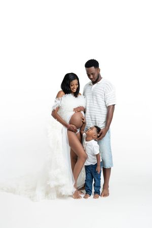 A pregnant woman in a white gown poses with her partner and young child in a bright studio maternity photo.