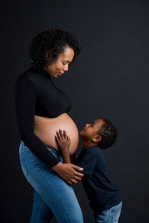 Pregnant Black woman in black top and jeans with young child touching her bare belly against dark background.