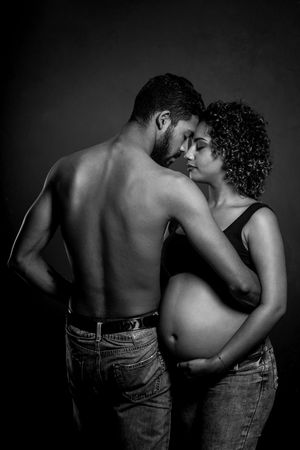 Black and white photo of a shirtless man embracing a pregnant woman in jeans, sharing an intimate moment.