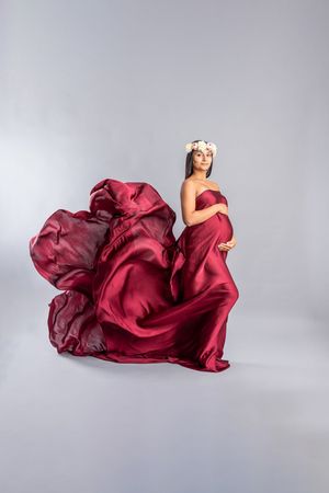 Pregnant woman draped in flowing red silk fabric wearing a floral crown, posing gracefully against a gray studio background.