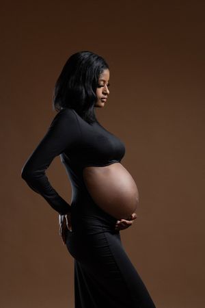 Pregnant Black woman in black dress cradling baby bump, side profile studio portrait on brown background.
