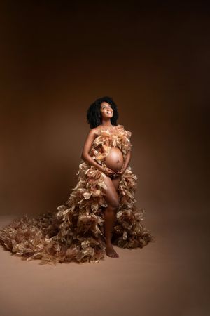 Pregnant Black woman in elegant ruffled tan gown cradling baby bump, posing gracefully against warm brown studio background.
