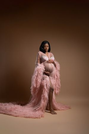 Pregnant woman in flowing pink tulle robe cradling baby bump in professional maternity studio portrait.