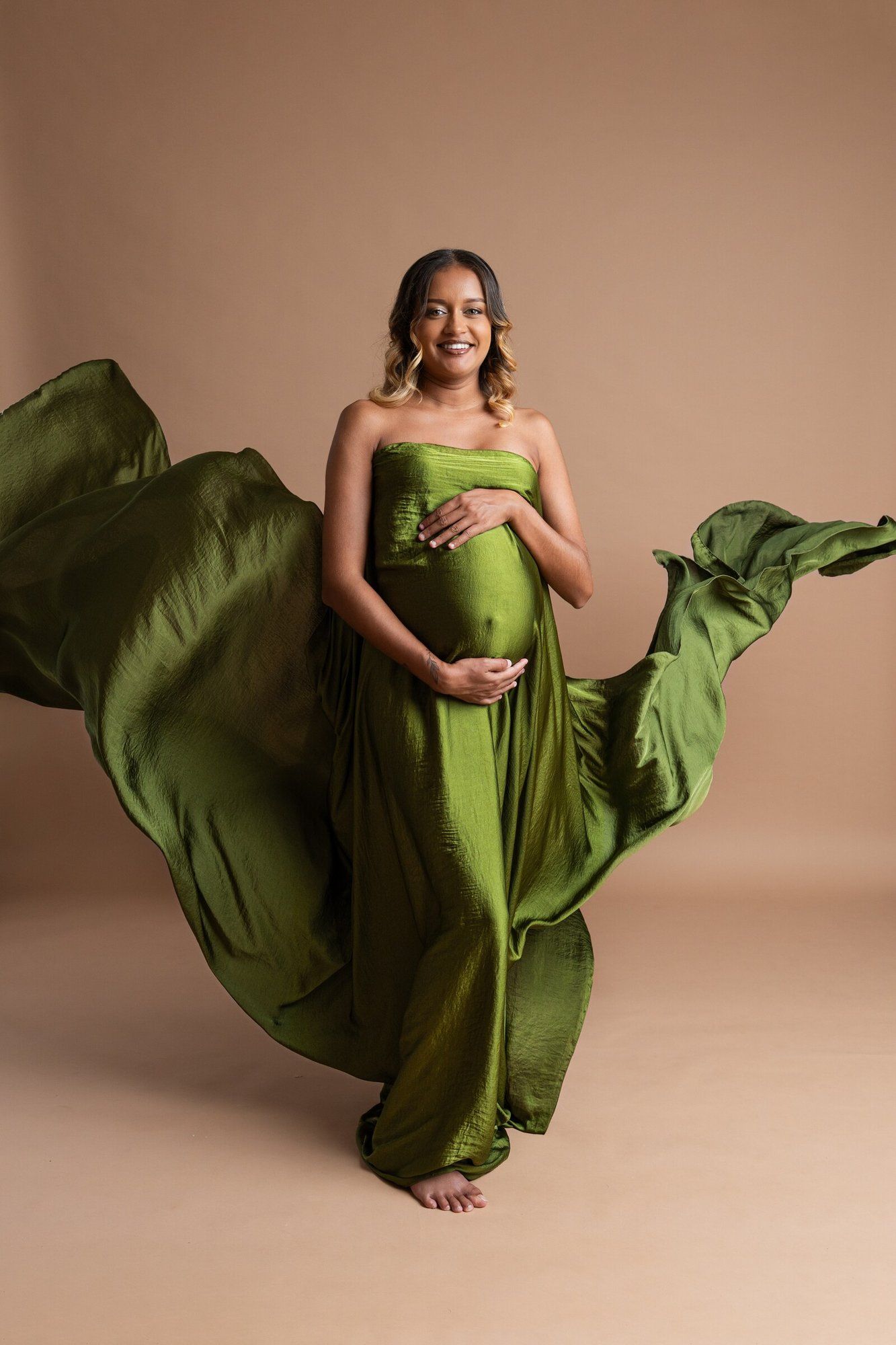 Pregnant woman in flowing olive green strapless gown with fabric billowing gracefully against beige background.
