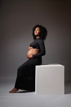 Smiling pregnant Black woman in black dress sitting on white cube, cradling baby bump in studio.