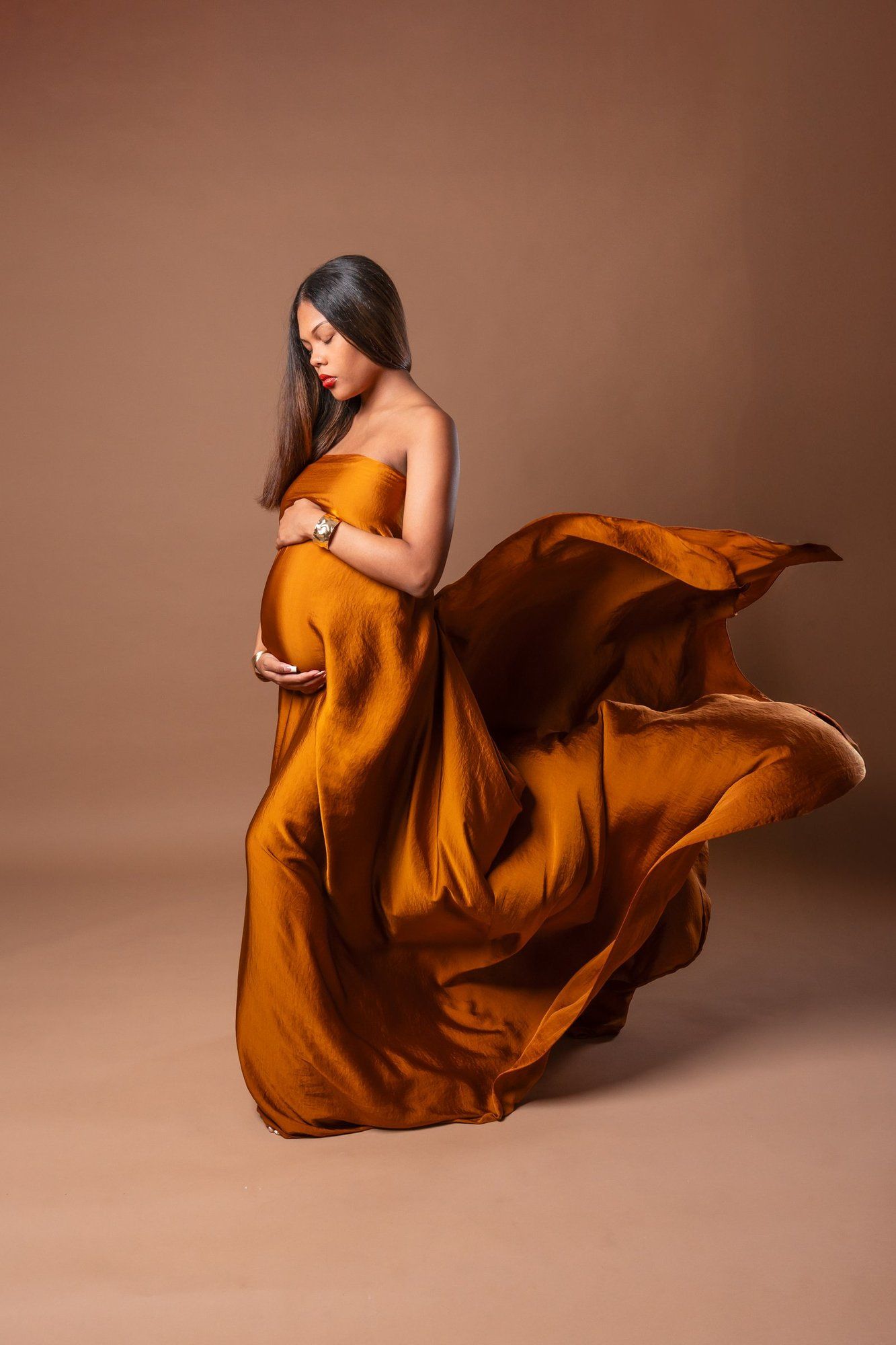 Pregnant woman in flowing burnt orange silk gown, elegantly draped, fabric billowing, studio portrait on warm beige background.