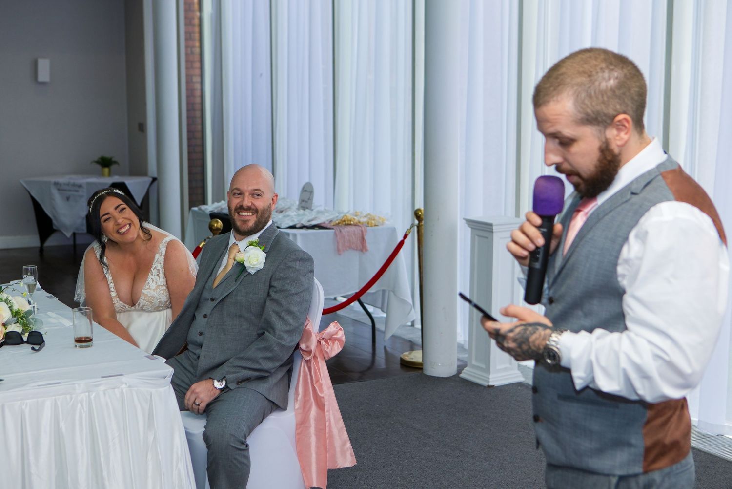 Best man gives a speech with purple microphone as bride and groom laugh at wedding reception.