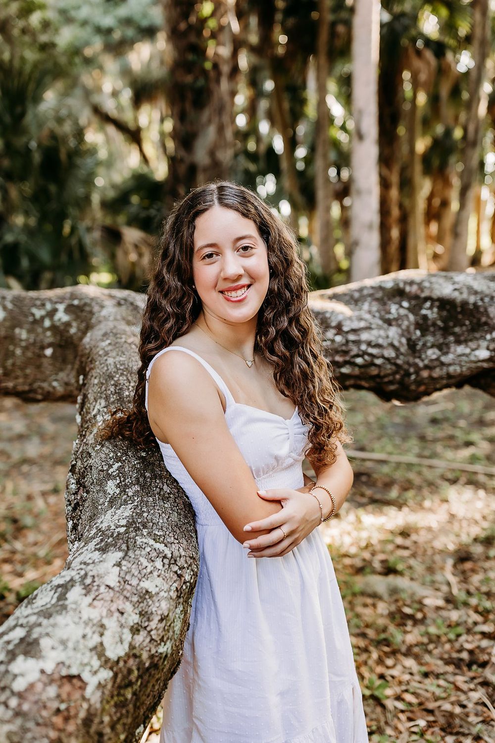 A smiling teenage girl in a white sundress leans against a mossy tree trunk in a sunlit forest setting.