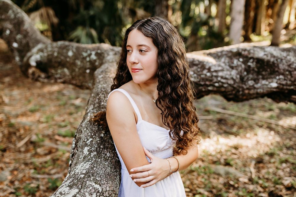 Young woman with curly hair in white dress leaning against mossy tree trunk in sunlit forest setting.