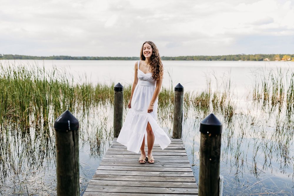 Young woman in white dress standing on a wooden dock by a serene lake on a cloudy day.