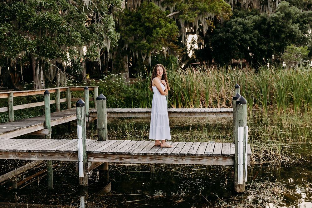 Young woman in white dress standing on wooden dock surrounded by lush greenery and calm water.