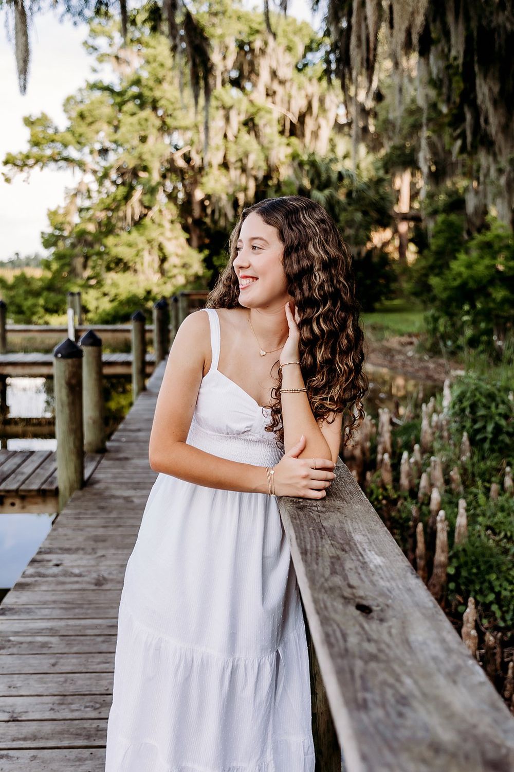 Young woman in white sundress smiling on wooden dock surrounded by lush green trees with Spanish moss.