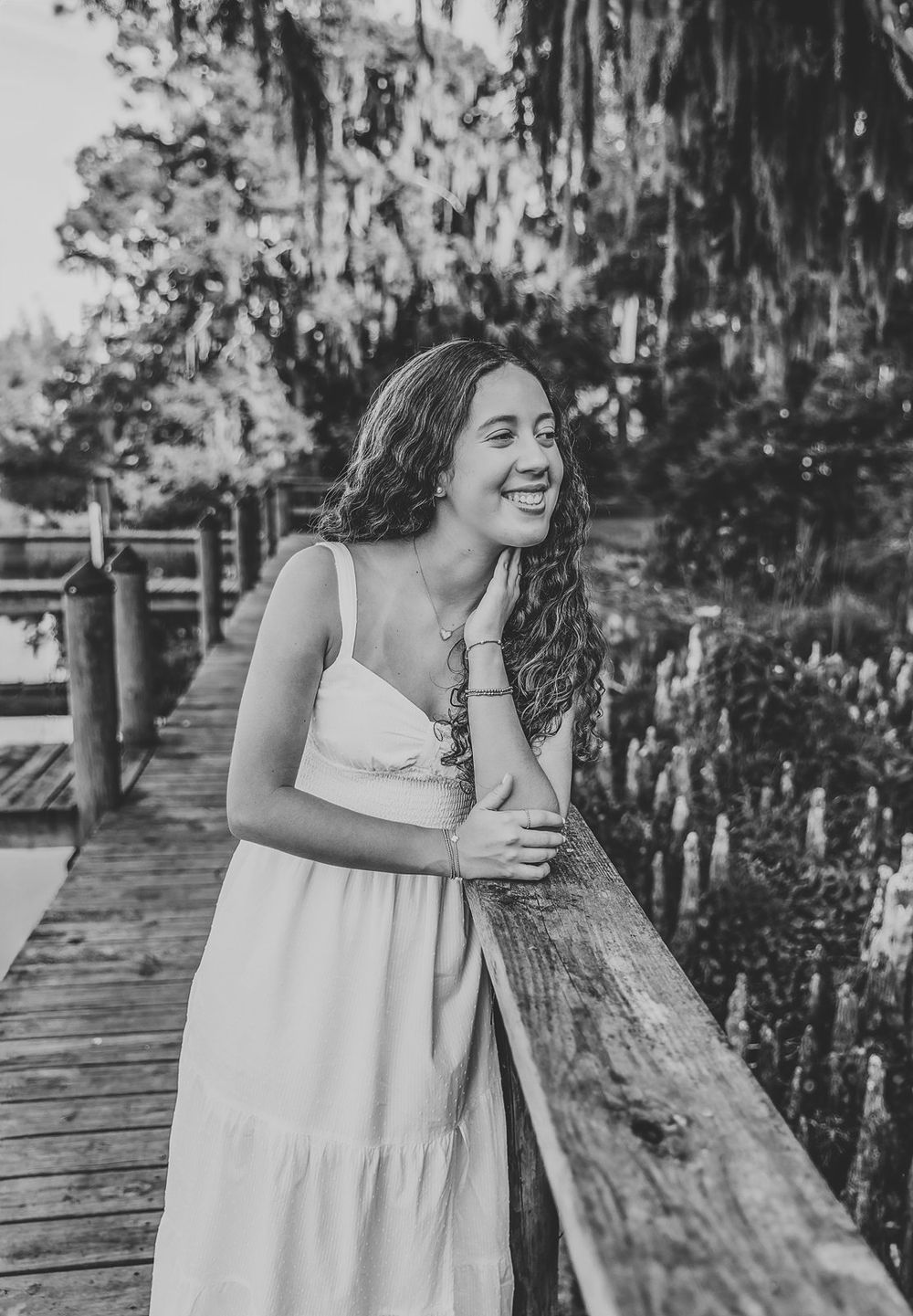 Young woman in white sundress smiling on wooden bridge surrounded by lush greenery, black and white photo.