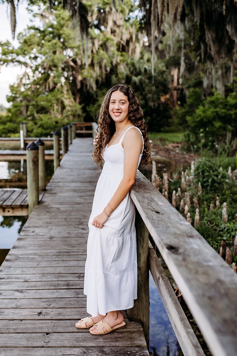 Young woman in white dress smiling on wooden dock surrounded by Spanish moss trees.