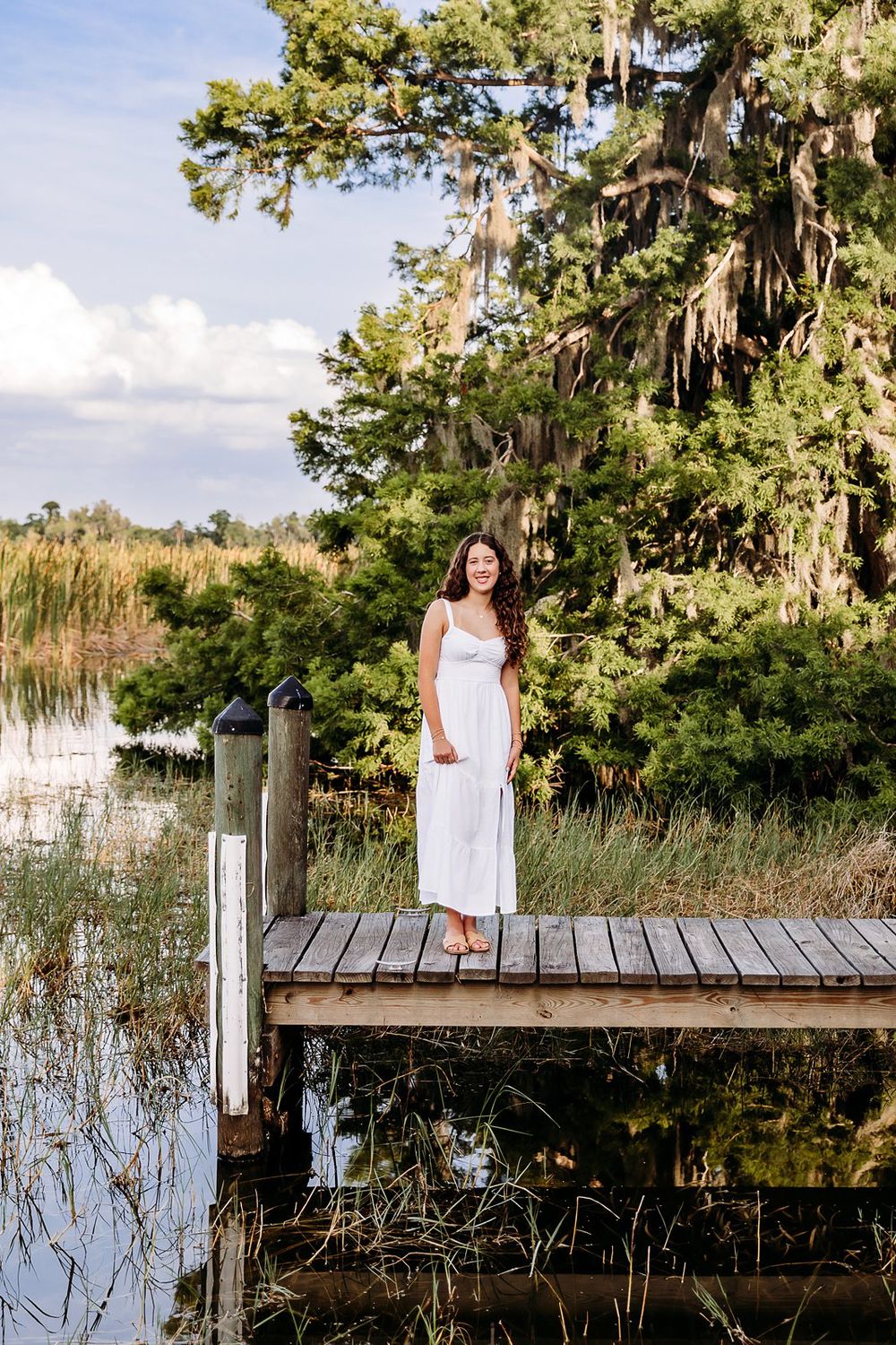 Young woman in white maxi dress standing on wooden dock surrounded by lush greenery and calm water.