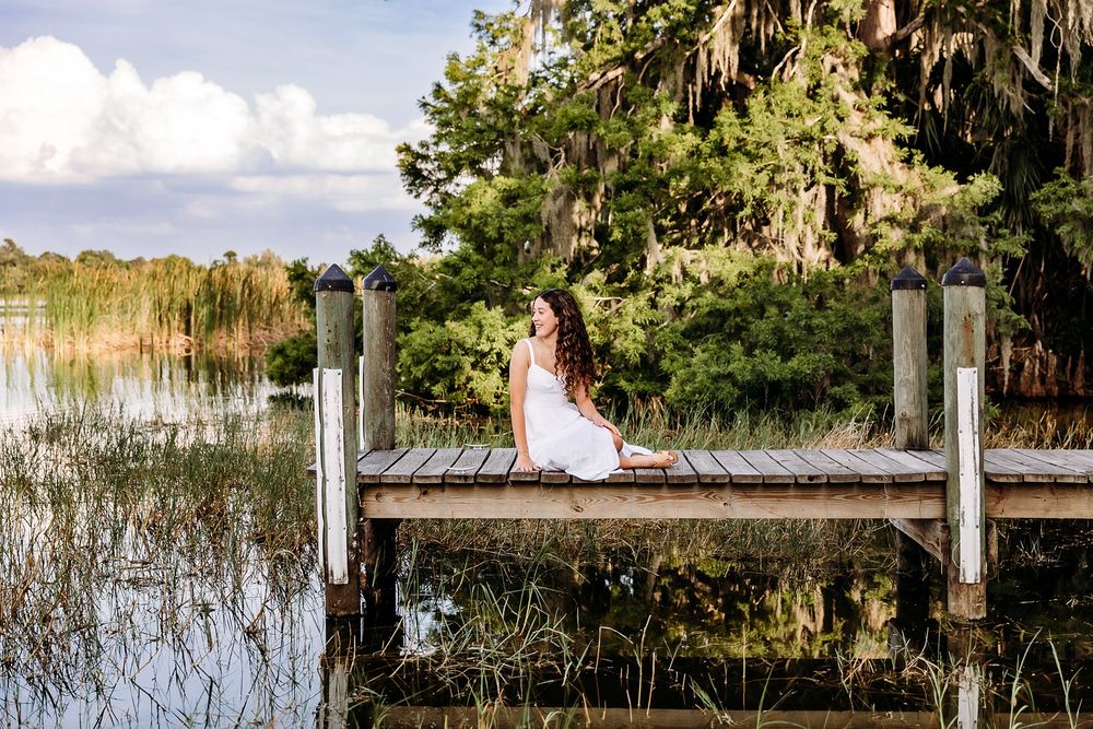 Young woman in white dress sitting on a wooden dock by a serene lake surrounded by lush green trees.