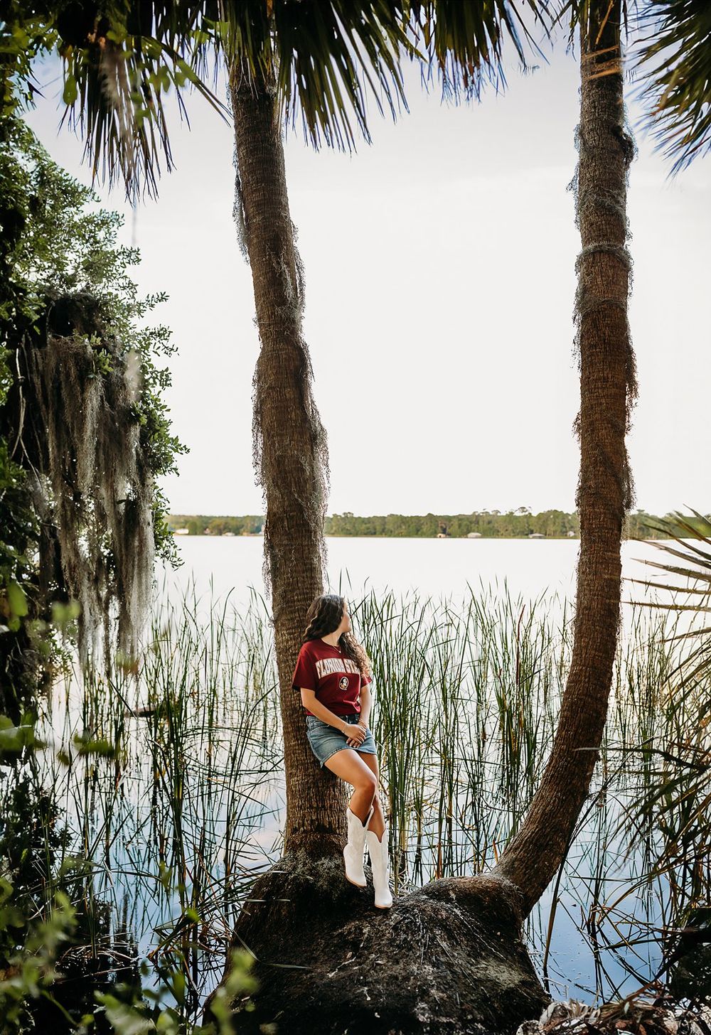 Young woman in maroon shirt and denim shorts poses between palm trees by a serene Florida lake with marsh grass.