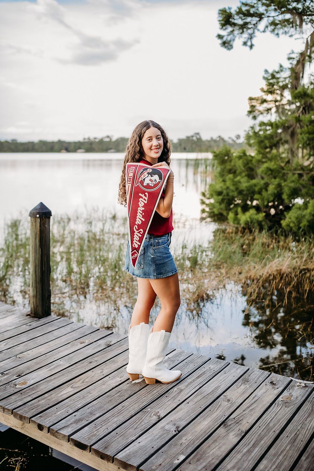 Young woman in white cowboy boots and denim skirt holds Florida State pennant on a wooden dock by a lake.