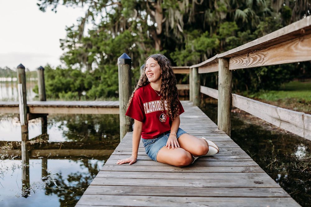Girl in Florida State shirt sitting cross-legged on wooden dock laughing by lake.