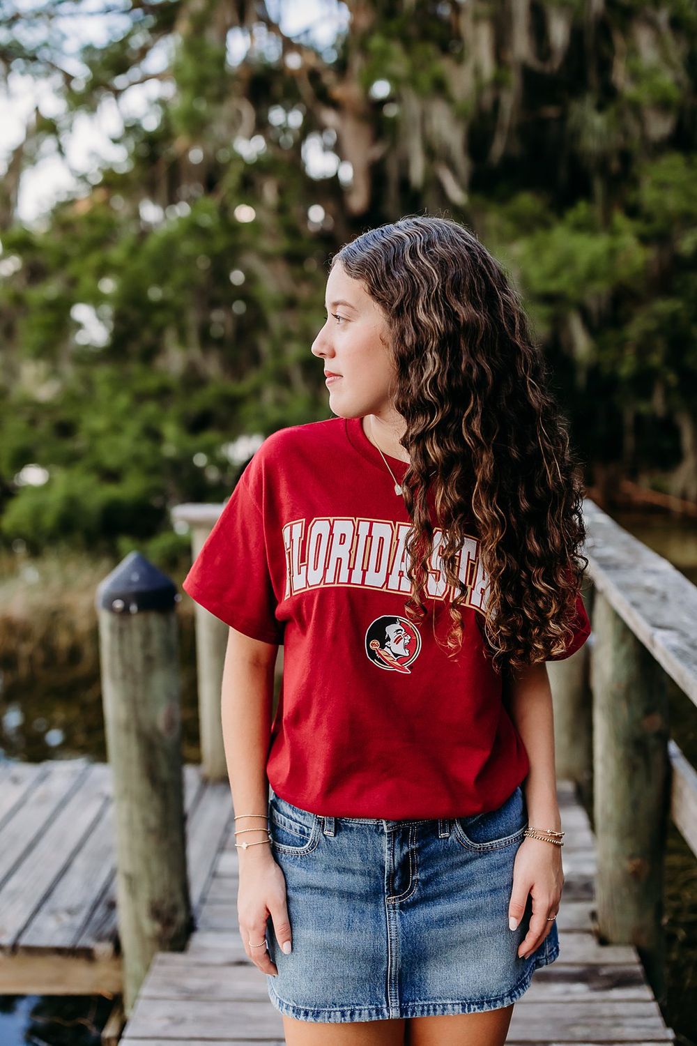 Young woman in Florida State red t-shirt and denim skirt standing on wooden dock surrounded by lush trees.