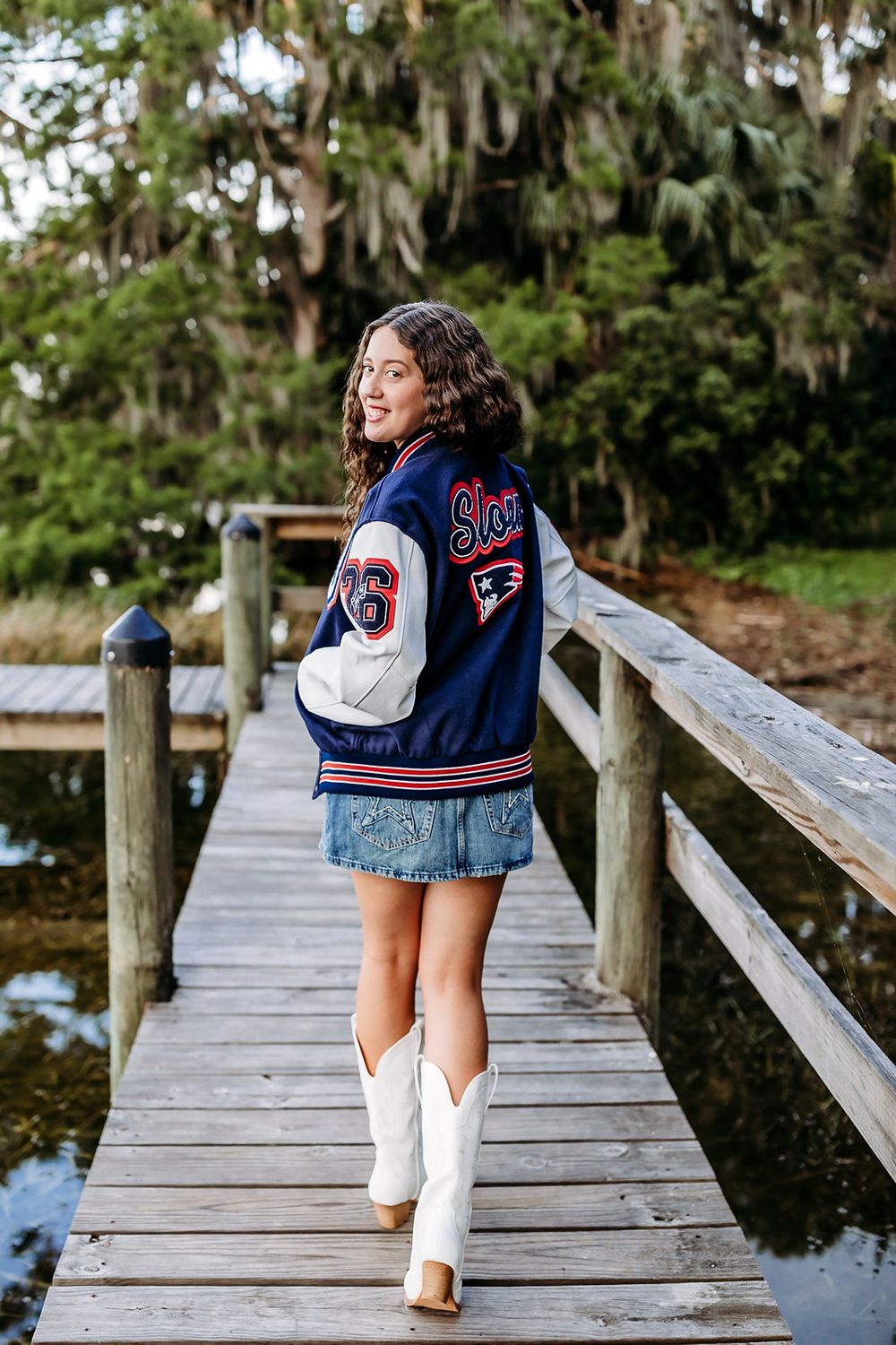 Teen girl in varsity jacket, denim skirt, and white cowboy boots posing on a wooden dock surrounded by trees.