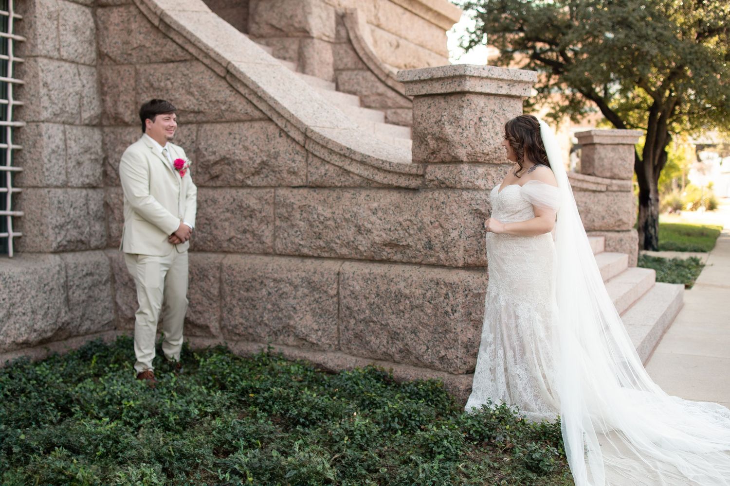 Bride in white lace gown and groom in tan suit share a first look moment by stone steps outdoors.